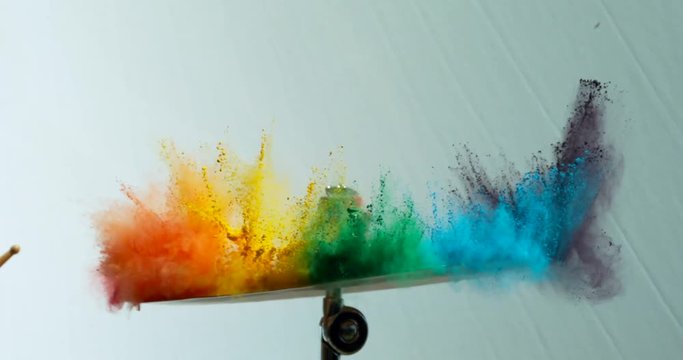 Colorful rainbow holi powder bounces off white canvas background in shockwave pattern of green blue pink and orange, slow motion, closeup