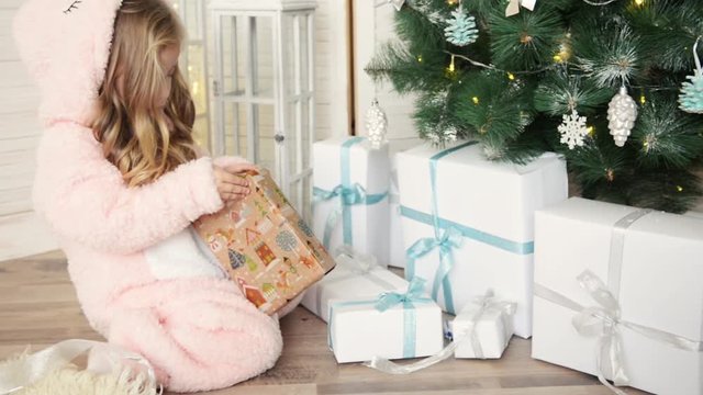 Happy Christmas little girl opening a wrapped Xmas gift box near decorated Christmas tree. Laughing child celebrating Christmas and New Year Winter Holidays.