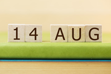 Wooden cube shape calendar for AUG 14 on green book, table.