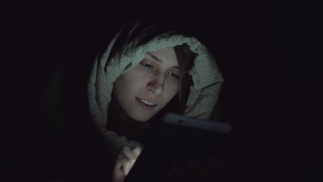 cute woman under a duvet in her bedroom staring at a glowing digital tablet screen at night, online chat, cinematic footage