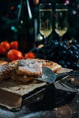 a festive New Year's table, cheese cake with chocolate, tangerines, grapes, biscuits, champagne and two glasses against the backdrop of a decorated Christmas tree with lights
