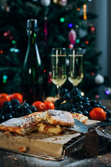 a festive New Year's table, cheese cake with chocolate, tangerines, grapes, biscuits, champagne and two glasses against the backdrop of a decorated Christmas tree with lights