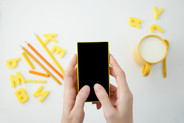 hands of a girl holding a smartphone, on a white table, yellow cup and yellow letters with pencils