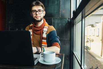 Handsome bearded male freelancer in glasses dressed in the warm sweater relaxing, drinking coffee and trying to get working with laptop near the big window in loft interior studio.