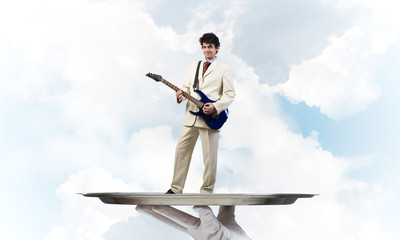 Fototapeta na wymiar Businessman on metal tray playing electric guitar against blue sky background