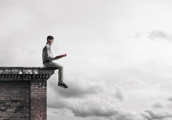 Man on roof edge reading book and cloudscape at background