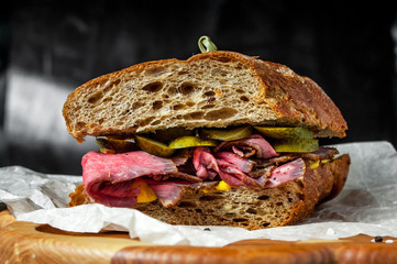 Pastrami sandwich on rye bread with pickles and mustard sauce, served on  wooden plate