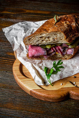 Pastrami sandwich on rye bread with pickles and mustard sauce, served on  wooden plate
