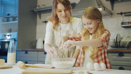 Cute pretty teen girl breaking an egg to the glass bowl with a daugh inside and her mother watching she doing this and controlling cooking. Portrait shot. - Powered by Adobe