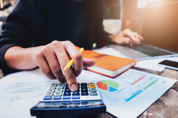 Business or accountant concept, man holding pen using calculator and laptop computer to analysys calculate financial & budget & tax data document with notebook in working on wood desk in office.