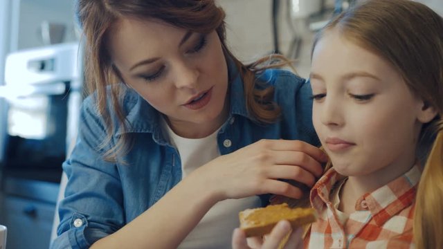 Portrait shot of the charming mother and cute daughter sitting and having rest while eating bread with peanut butter and drinking juice and tea. Close up. Indoor