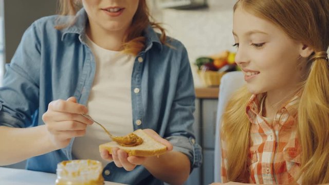 Young attractive mother spreading a peanut butter on the bread and talking with her teen daughter while they sitting at the table at the kitchen. Close up. Inside