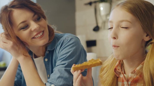 Close up of the cute pretty teen girl enjoying a sandwich with peanut butter and her mother looking at her, than hugging and kissing her. Portrait shot. Indoors