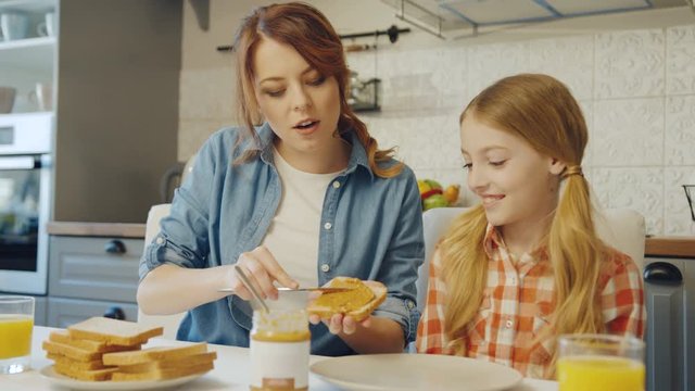 Beautiful mother showing to her pretty blonde teen girl how to spread the peanut butter on the bread in the nice cozy kitchen. Portrait shot. Inside