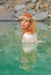 A beautiful red-headed girl with flowers  in her hair is dressed in vintage clothing. She relaxes in a quarry pond filled with  green water.