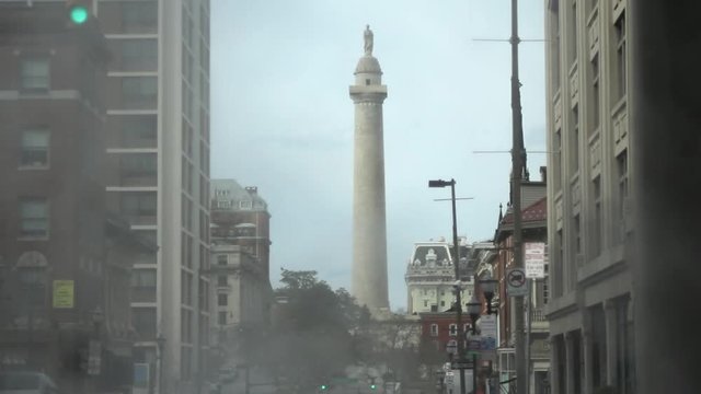 Medium shot of steam and traffic passing in front of the 178 foot doric column Washington Monument in the Mount Vernon neighborhood.