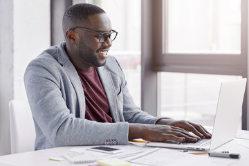 Pleased black Afro American male enterpreneur checks online database of finance report on laptop computer, uses free internet connection. Dark skinned man types messages to friends in social networks