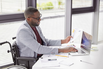 Smiling dark skinned young businessman sits in wheelchair, being disabled or invalid, develops business project, has happy expression, keyboards on laptop computer, makes statistics and graphics