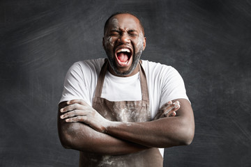 Happy overjoyed black male cook keeps hands crossed, laughs loudly at good joke of his colleague,...