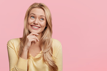 Dreamful pretty blonde woman with long hair has appealing smile, looks thoughtfully aside, poses...