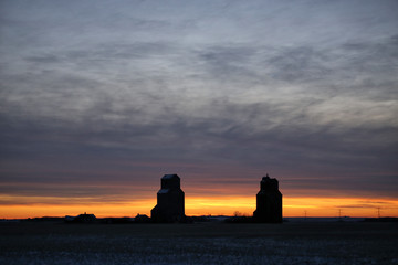Fototapeta premium Two old wooden grain elevators silhouetted under a sunset sky in a countryside winter landscape