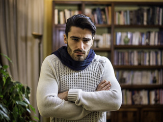 Young man with scarf, holding his arms crosssed for coldness, at home in his living room