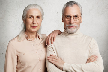 Glad grey haired wrinkled woman being pleased to meet her first love, leans at senior man`s shoulder, remember pleasant moments of their relatioship. Senior couple stand next to each other indoor