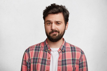 Photo of handsome young man with trendy hairdo and mustache, curves lips and frowns face in bewildermennt, isolated over white concrete background. Attractive male student in casual clothes.