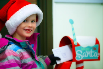 A five year old in a santa hat with a big smile putting a letter into a red Christmas post box for santa