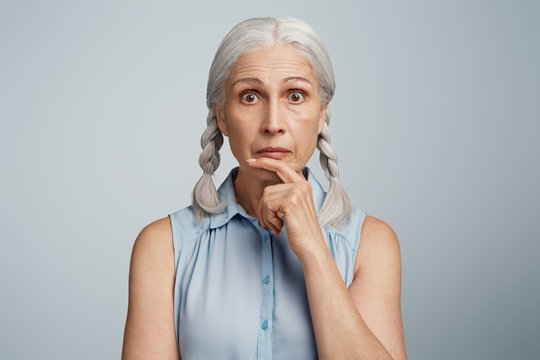 Mature shocked female has two pigtails, looks with surprised expression directly at camera, being amased by news which she heard on TV. Attractive senior woman poses in studio against blank wall