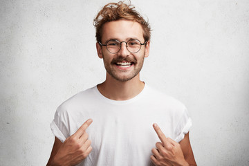 Smiling young male indicates at blank t shirt, has happy expression, glad to advertize new clothes,...