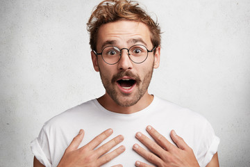 Amazed young stylish guy keeps hands on chest, opens mouth in bewilderment, stares at camera,...