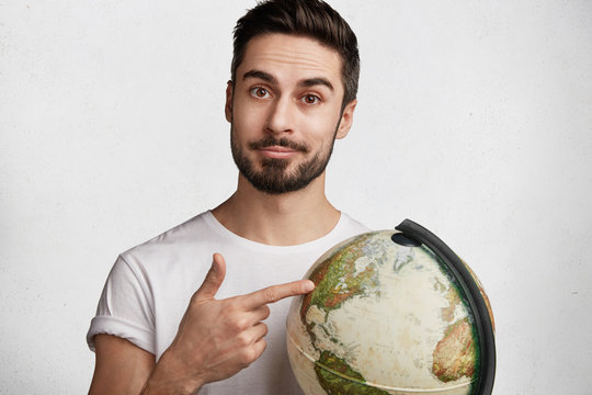 Portrait of serious confident unshaven man with trendy hairstyle, holds globe, chosses destination for travelling with girlfriend, likes to explore new unknown places, isolated over white background