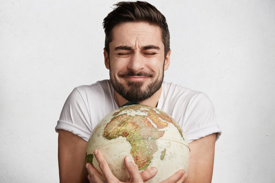 This is my palnet! Pleased emotional bearded young male closes eyes with enjoyment as embraces globe, expresses love to whole planet and world, likes travelling. People, tourism, emotions concept