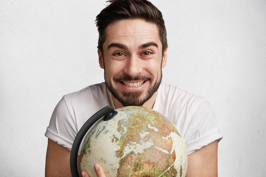 Indoor shot of unsahven tourist embraces globe with cheerful expression, demonsrtates his love to travelling, happy to explore many countries and their traditions, isolated over white concrete wall