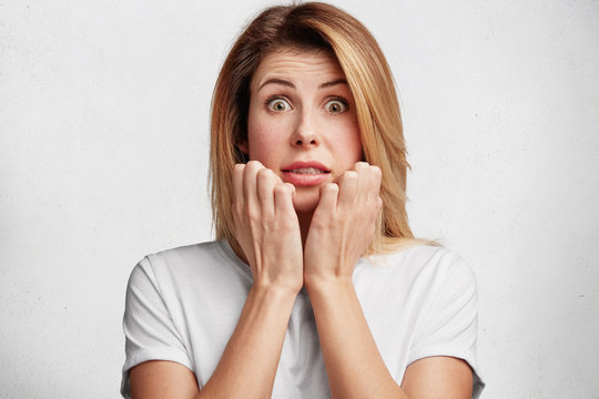 Surprised emotional female model with luxurious hair, clenches teeth and trembles in terror, afriad of being asked on public eye, worries because of some importnat events, isolated on white wall