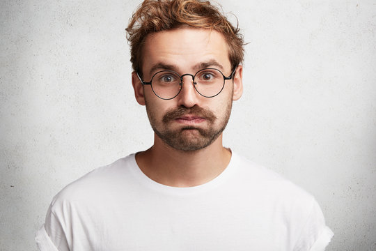 Portrait of tired stylish bearded intelligent male student gives deep sigh, presses lips, feels relief after passed exam, isolated over white background. Handsome young unshaven man poses indoor.