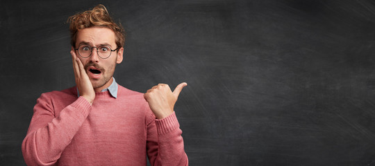 Horizontal shot of amazed shocked displeased fashionable male indicates at something with surprised expression, poses against black chalk background, copy area for promotional content or text