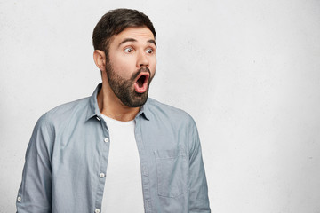 Stupefied attractive brunet male wears shirt, stares with great surprisment aside, looks at watch, realizes that he is late to catch transport, isolated over white concrete background with copy space