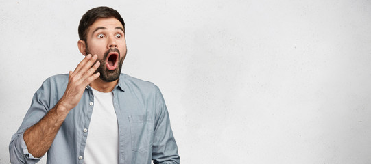 Horizontal studio shot of shocked frightened male model has hush reaction as sees his phobia, keeps mouth opened, dressed casually, isolated over white background with copy space for your text