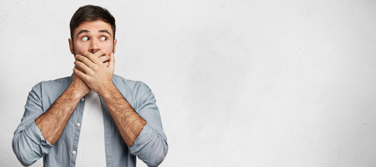 I will not say anything! Young male model has dark hair covers mouth with hands, dressed casually,...