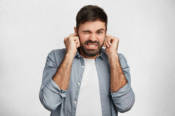 Handsome bearded young male model plugs ears with displeased expression as hears annoying loud...