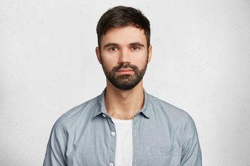 Obraz premium Portrait of confident serious male model has black beard and mustache, dressed in casual denim shirt, looks seriously into camera, isolated over white background. Hipster guy models in white studio