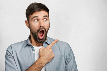 Horizontal portrait of shocked young male has thick beard and mustache looks with widely opened...