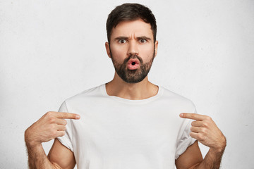 Amazed strict shocked young bearded male model poses against white studio background, dressed in...