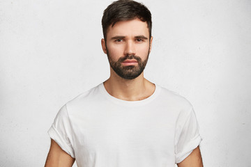 Horizontal portrait of unshaven stylish young man dressed in white new t shirt, looks seriously directly in camera, expresses his confidence, rests at home after hard working day, isolated on white