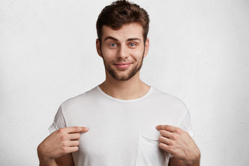 Positive glad blue eyed attractive man with stubble indicates at blank space of t shirt for your advertising text or logo, isolated over white background. People, clothing, design and fashion concept