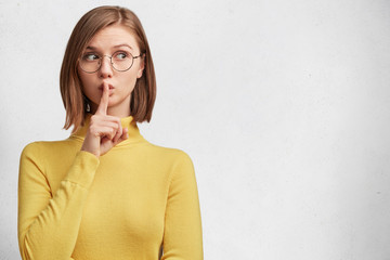 Horizontal shot of cute lovely woman keeps fore finger on lips, looks silently aside, wears casual...