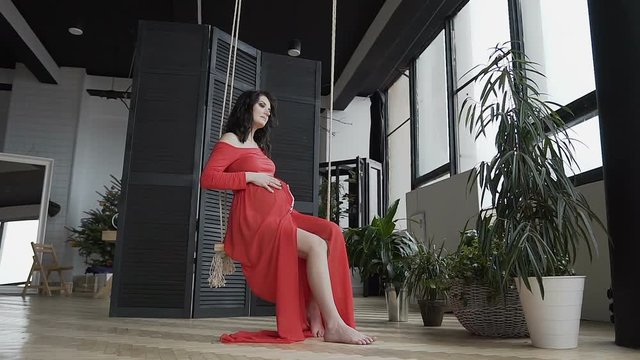 A pregnant girl in a long red dress sits on the swing and gently strokes her belly and looks at him. Conception of pregnancy. Happy time