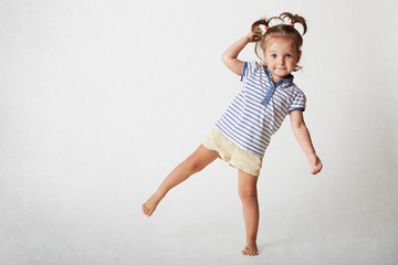 Small adorable female child has funny expression, two pony tails, wears casaul t shirt and shorts, stands on one leg, poses against white background. Playful little kid glad to be photographed